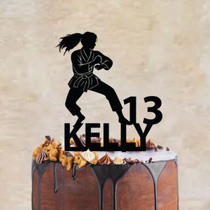 Peut inclure: Un gâteau topper noir représentant la silhouette d'une personne en uniforme d'arts martiaux effectuant un coup de pied, placé au-dessus du nom KELLY et du chiffre 13 dans une police grasse, le tout formant une seule pièce décorative.
