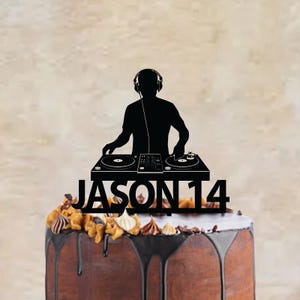 Op de afbeelding: Een zwarte taarttopper met een silhouet van een DJ die een koptelefoon draagt en een draaitafelconsole bedient. Onder de DJ staat de tekst JASON 14 in een vet, schreefloos lettertype. De topper is geplaatst bovenop een chocoladetaart.