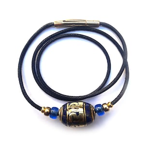 Puede incluir: Un collar de cordón negro con una cuenta dorada y azul. La cuenta tiene letras doradas.