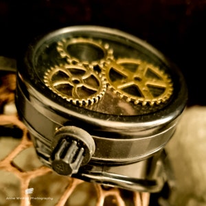 Könnte beinhalten: Nahaufnahme eines schwarzen runden Objekts mit einer durchsichtigen Glasoberfläche. Unter dem Glas befinden sich drei goldene Zahnräder, ein großes und zwei kleinere, die ineinandergreifen und sichtbar sind.