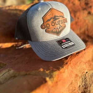 May include: A gray and black trucker hat with the words "GO OUTSIDE" on a brown leather patch. The patch features a mountain and tree design. The hat has a curved brim and an adjustable strap.