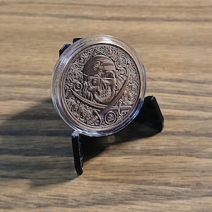 May include: A bronze-colored coin featuring a pirate skull with crossed swords, displayed in a clear protective case. The coin is set on a black stand against a wooden background. The pirate design includes a bandana and intricate details.