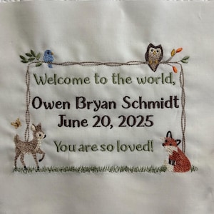 May include: Embroidered white fabric with the text "Welcome to the world, Owen Bryan Schmidt June 20, 2025 You are so loved!" framed by a brown border. The design includes a blue bird, owl, deer, fox, and butterfly.