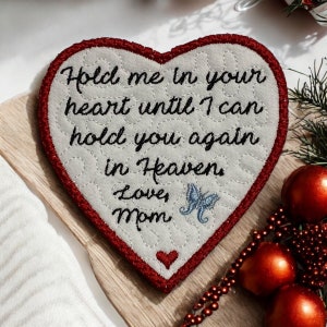 Peut inclure: Une décoration en tissu en forme de cœur blanc avec des coutures rouges autour des bords. L'ornement a un message brodé qui dit "Hold me in your heart until I can hold you again in Heaven. Love, Mom" et un petit papillon bleu.