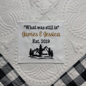 May include: A white fabric square with black and gold embroidery featuring the text "What was still is", "James & Jessica", and "Est. 2019" above a black embroidered mountain and pine tree landscape, stitched onto a white quilted fabric background.