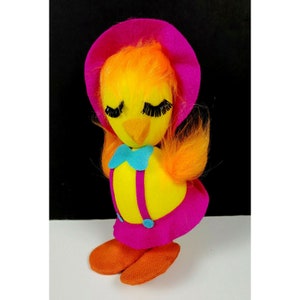 May include: A yellow plush bird with orange hair, pink and white dress, and blue bow. The bird has closed eyes with long eyelashes and a pink hat.