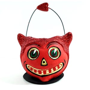 May include: A red devil-faced Halloween candy bucket with glitter accents on the horns and rim. The bucket has a black handle and a heart-shaped accent. The face has green eyes, a triangle nose, and a smiling mouth. The bucket sits on a black base.