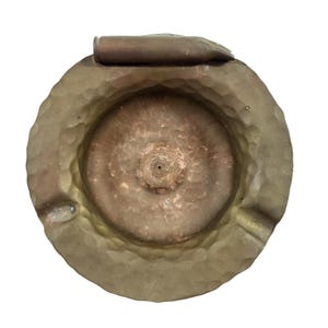 May include: A circular, hammered-texture metal ashtray with a dull, brownish-gold finish. A single spent rifle cartridge casing is positioned horizontally across the top rim of the ashtray, which features a central circular indentation.