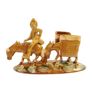 May include: A glazed ceramic figurine featuring a person riding a donkey, followed by a second donkey carrying a large basket. The piece is finished in shades of brown and tan, set upon an oval base with green and brown painted accents.