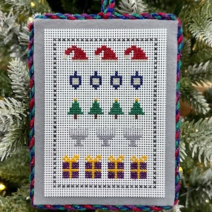 May include: A grey felt Christmas bauble with a white cross-stitch design featuring Santa hats, baubles, Christmas trees, presents, and reindeer. The bauble is trimmed with a red, blue and green braided cord.