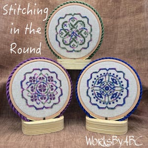 Puede incluir: Tres aros de bordado con diseños de bordado morado, verde y azul. Los aros están en soportes de madera y los diseños son todos diferentes. El texto "Stitching in the Round" está en la parte superior de la imagen.