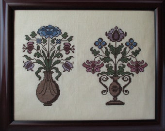 May include: Two embroidered floral arrangements in brown, pink, blue, and green tones, each in a brown vase, framed in a dark brown frame.