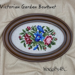 May include: Oval framed cross-stitch of a floral bouquet. The bouquet features blue, red, and pink flowers with green leaves. The frame is a dark brown color. Text at the top reads "Victorian Garden Bouquet" and text at the bottom reads "WorksByABC".