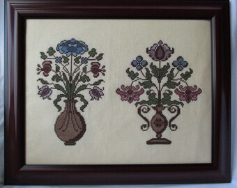 May include: Two framed cross-stitch embroidery pieces featuring floral arrangements in vases. The left piece has blue, pink, and green flowers in a brown vase. The right piece has pink, blue, and green flowers in a brown vase with a decorative base.