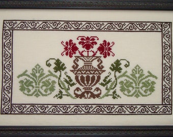 May include: A framed cross-stitch embroidery featuring a stylized floral design with a vase in the center. The vase is brown and white, and the flowers are red and green. The embroidery is surrounded by a black and white border.