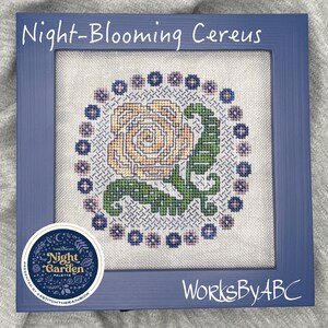 Peut inclure: Un motif de point de croix encadré représentant une fleur de cactus à fleurs nocturnes avec une tige et des feuilles vertes. La fleur est entourée d'un cercle de petites fleurs violettes et bleues. Le cadre est de couleur bleu foncé et porte le texte "Night-Blooming Cereus" en haut et "WorksByABC" en bas.