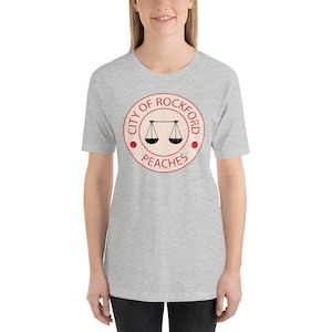 Op de afbeelding: Grijs T-shirt met een rode cirkelvormige afbeelding met de tekst "City of Rockford Peaches" en een weegschaalontwerp in de cirkel.