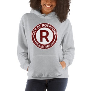 Op de afbeelding: Een lichtgrijze hoodie met een voorzak en een capuchon met trekkoord. De hoodie heeft een cirkelvormige afbeelding met de tekst "CITY OF ROCKFORD" bovenaan, "PEACHES" onderaan en een grote "R" in het midden.