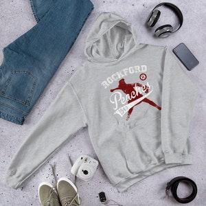 Pode incluir: Camisola de capuz cinzenta com um gráfico vintage vermelho de um jogador de beisebol e o texto "Rockford Peaches 1943" com um símbolo de marca registada.