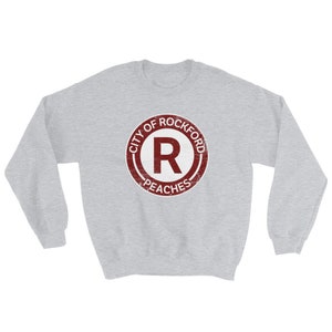 Op de afbeelding: Een lichtgrijs sweatshirt met een cirkelvormige bordeauxrode afbeelding. De afbeelding bevat de tekst "CITY OF ROCKFORD" bovenaan, een grote "R" in het midden en "PEACHES" onderaan. Het ontwerp heeft een vintage uitstraling.