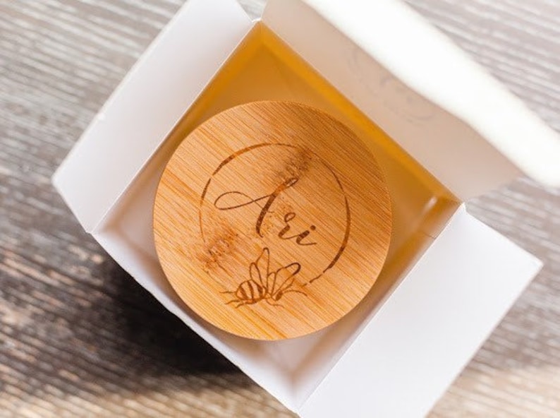 May include: A wooden lid with the name "Ari" engraved on it. The lid has a bee design and is in a white box.