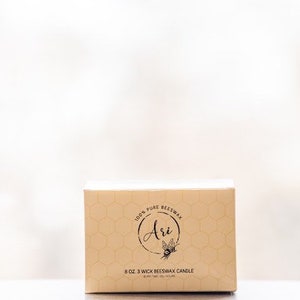 May include: A beige box with a honeycomb pattern and the text "100% Pure Beeswax" and "Ari" with a bee illustration. The box also says "8 oz 3 Wick Beeswax Candle".