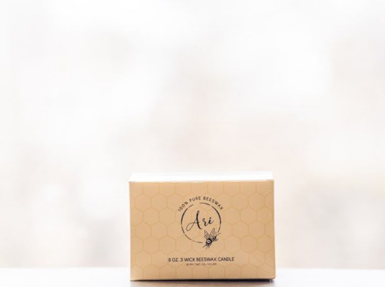 May include: A beige box with a honeycomb pattern and the text "100% Pure Beeswax" and "Ari" with a bee illustration. The box also says "8 oz 3 Wick Beeswax Candle".