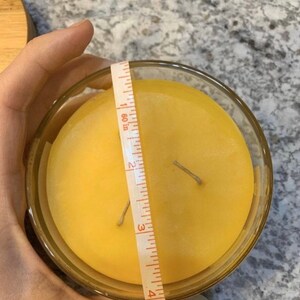 May include: A yellow candle in a clear glass jar with two wicks. The candle is being measured with a tape measure that reads 4 inches.
