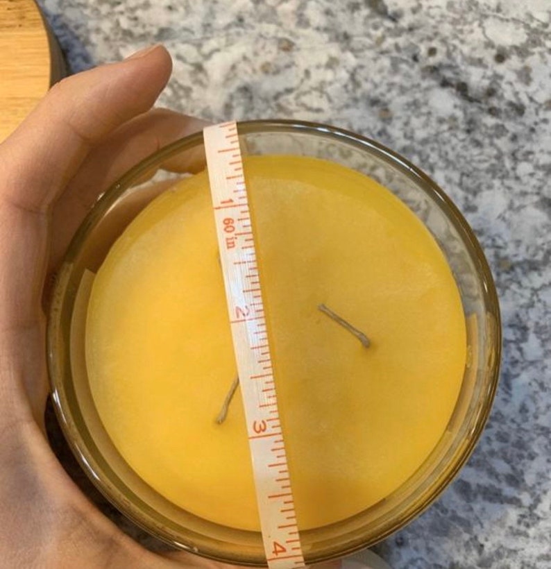 May include: A yellow candle in a clear glass jar with two wicks. The candle is being measured with a tape measure that reads 4 inches.