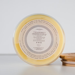 May include: Close-up of the bottom of a glass jar candle with a yellow wax base. The label on the jar reads "WARNING" and includes information about the candle's ingredients and usage. The jar has a bamboo lid.