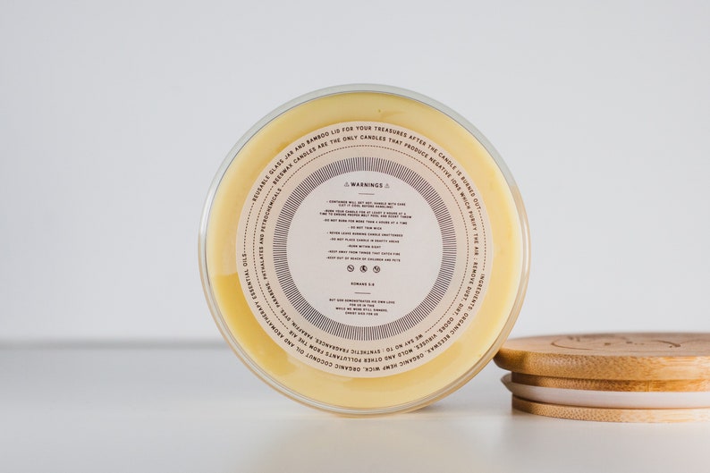 May include: Close-up of the bottom of a glass jar candle with a yellow wax base. The label on the jar reads "WARNING" and includes information about the candle's ingredients and usage. The jar has a bamboo lid.