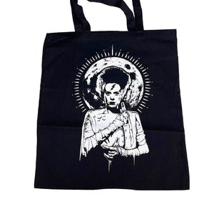 以下が含まれることがあります： 白いステッチのついたモンスターが満月の前に立っている、白いグラフィックが描かれた黒いトートバッグ。