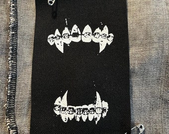 May include: A rectangular black fabric patch featuring a white screen-printed graphic of upper and lower teeth with metal braces. The patch is attached to a grey textured garment with two silver-toned metal safety pins at the top and bottom corners.