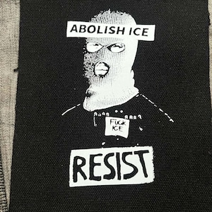May include: A rectangular black fabric patch featuring a white screen-printed graphic of a person wearing a balaclava. The text ABOLISH ICE appears over the eyes, FUCK ICE on a small square, and RESIST at the bottom. The patch is sewn onto tan fabric.