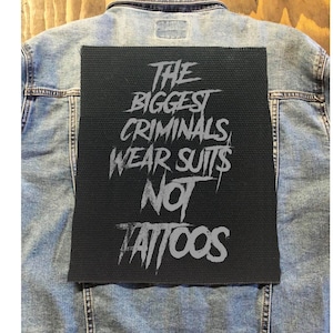May include: A black denim jacket with a gray patch on the back that says "The Biggest Criminals Wear Suits Not Tattoos".