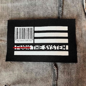 Puede incluir: Parche blanco y negro con un código de barras, tres rayas blancas y el texto "F**K THE SYSTEM" con una línea roja sobre la primera palabra. El parche está hecho de una tela negra.