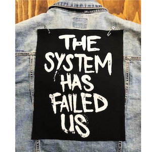 May include: Black denim jacket with a white patch sewn on the back. The patch says "THE SYSTEM HAS FAILED US" in white paint.