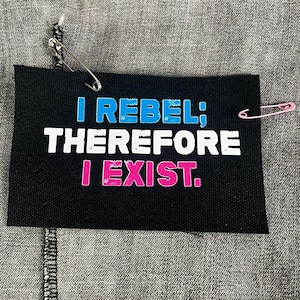 May include: Black rectangular patch with the text "I REBEL; THEREFORE I EXIST." in white, blue, and pink. The patch is attached to gray denim with safety pins.