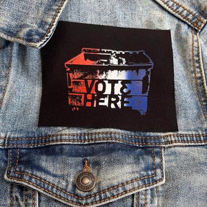 Könnte beinhalten: Ein schwarzer Patch mit einem weißen Müllcontainer mit roten, weißen und blauen Farbschleudern. Der Text "VOTE HERE" ist in Weiß auf den Müllcontainer gedruckt.