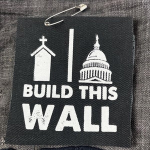 May include: Black and white patch with a church, a building, and the text "BUILD THIS WALL". The patch is attached to a gray fabric with a safety pin.