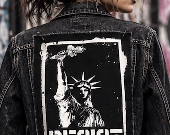 Puede incluir: Una chaqueta de mezclilla negra con efecto lavado ácido que presenta un gran parche rectangular en la espalda. El parche muestra un gráfico blanco de alto contraste de la Estatua de la Libertad con una antorcha y el texto RESIST.
