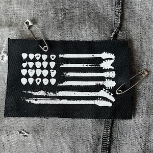 May include: A black and white guitar pick patch with an American flag design. The patch is pinned to a grey denim jacket with safety pins.