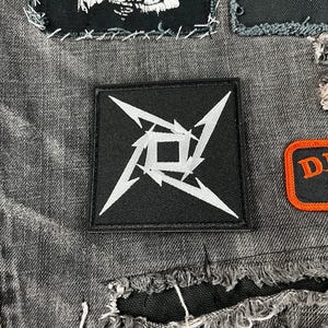 May include: A black square patch featuring a white, stylized, four-pointed star design. The patch is sewn onto a grey denim jacket, alongside other patches with band logos and graphics. The jacket has distressed details.