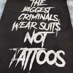 May include: A rectangular black fabric patch featuring the white printed text THE BIGGEST CRIMINALS WEAR SUITS NOT TATTOOS in a distressed, jagged font. The patch is sewn onto a light blue denim garment, with another partial black patch visible above.
