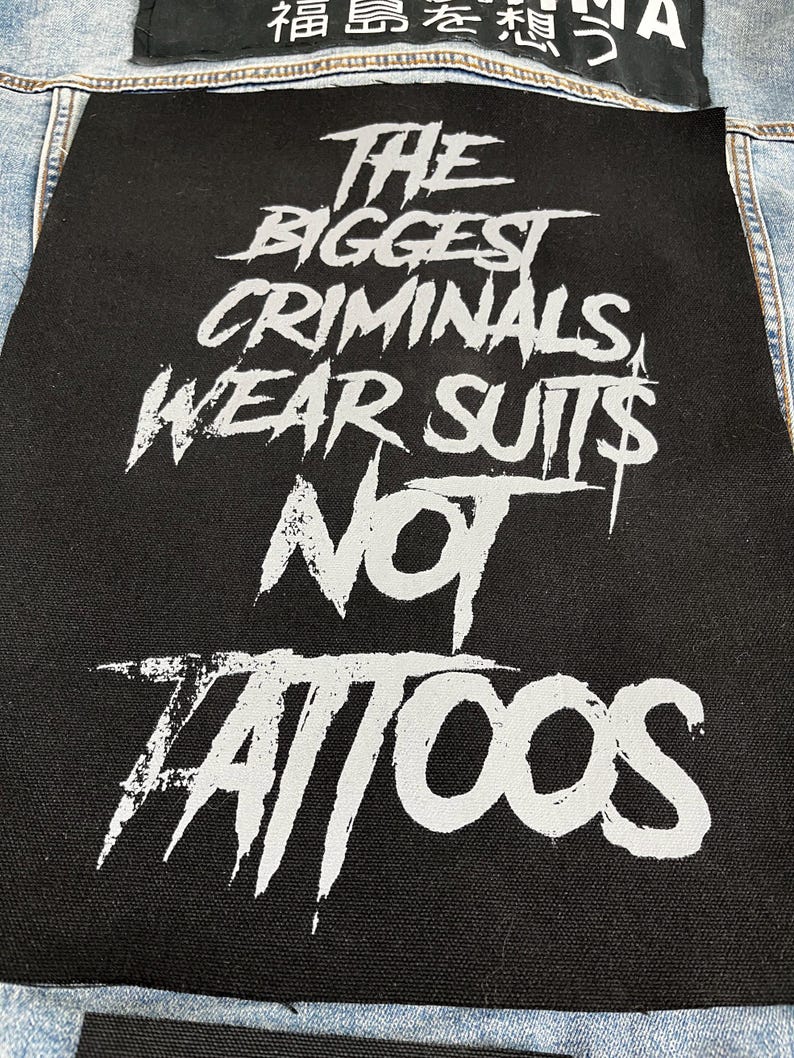 May include: A rectangular black fabric patch featuring the white printed text THE BIGGEST CRIMINALS WEAR SUITS NOT TATTOOS in a distressed, jagged font. The patch is sewn onto a light blue denim garment, with another partial black patch visible above.