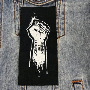 May include: A black rectangular patch featuring a white fist graphic and the text "SMASH THE OLIGARCHY". The patch is attached to a light blue denim jacket.
