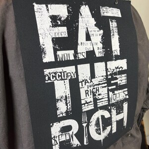 Pu&ograve; includere: Una toppa nera e bianca con il testo "EAT THE RICH" in grassetto in lettere bianche. Il testo &egrave; sovrapposto ad altro testo, tra cui "IT'S WOOT'S FOR SUPPER", "OCCUPY", "TAX THE RICH" e "$FTW".