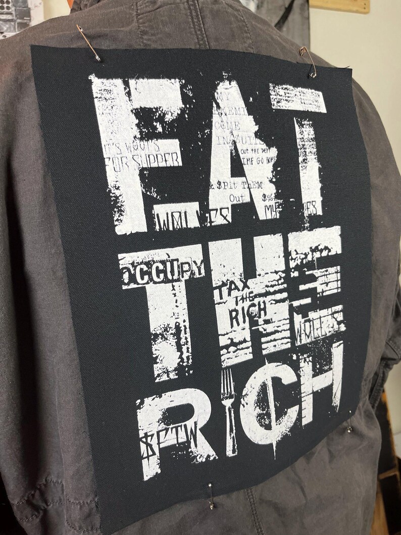 Pu&ograve; includere: Una toppa nera e bianca con il testo "EAT THE RICH" in grassetto in lettere bianche. Il testo &egrave; sovrapposto ad altro testo, tra cui "IT'S WOOT'S FOR SUPPER", "OCCUPY", "TAX THE RICH" e "$FTW".