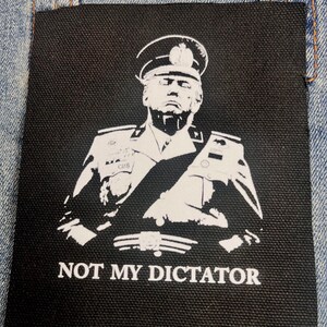 Op de afbeelding: Een vierkante zwarte stoffen patch met een witte zeefdruk van een persoon in een uniform en pet in militaire stijl. Onder de figuur staat de tekst NOT MY DICTATOR in een vetgedrukt, wit, hoofdletter serif-lettertype op de zwarte achtergrond.
