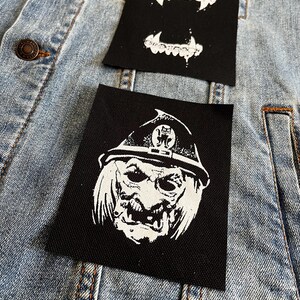 Peut inclure: Deux patchs thermocollants sur une veste en jean bleue. Un patch est noir avec une image blanche d'un monstre portant un chapeau et une grande bouche. L'autre patch est noir avec une image blanche d'une paire de crocs.